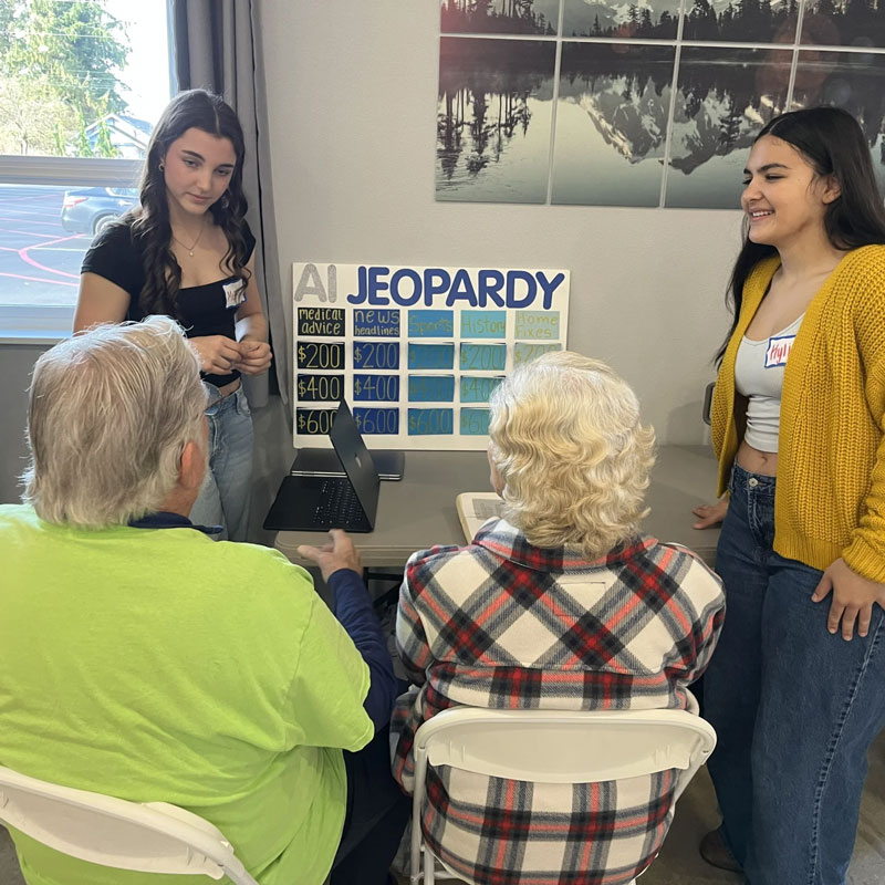Two teems stand next to an poster board titled AI Jeopardy with 2 seniors in folding chairs playing the game.